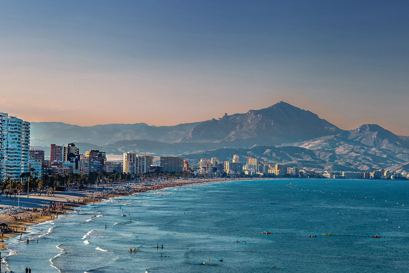 Panoramic view of Alicante's waterfront, historic castle, and city buildings under blue sky.