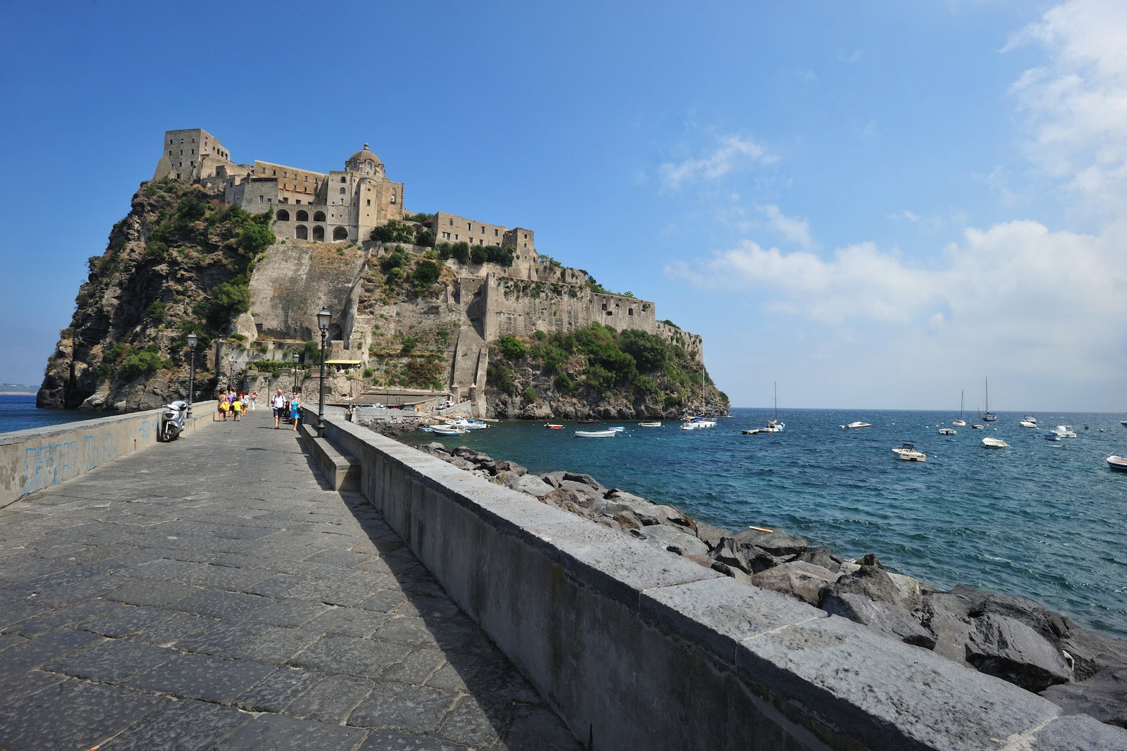 Historic Castel Nuovo castle in Naples, Italy, with boats in the harbor and Mount Vesuvius in the background.