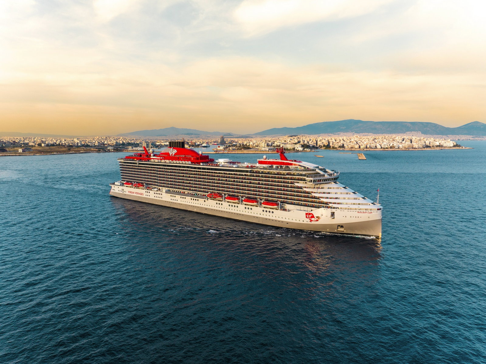 A modern cruise ship with a red funnel sailing on clear blue waters under a sunny sky.